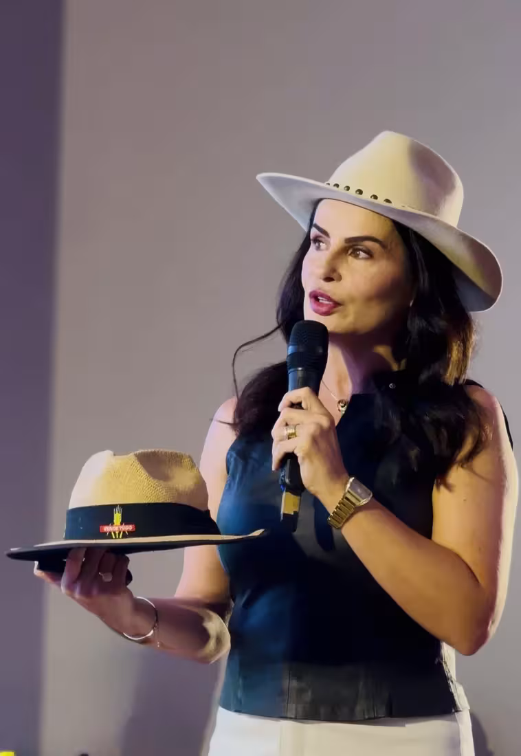 Mara Lauxen falando ao microfone e segurando um chapéu durante uma palestra